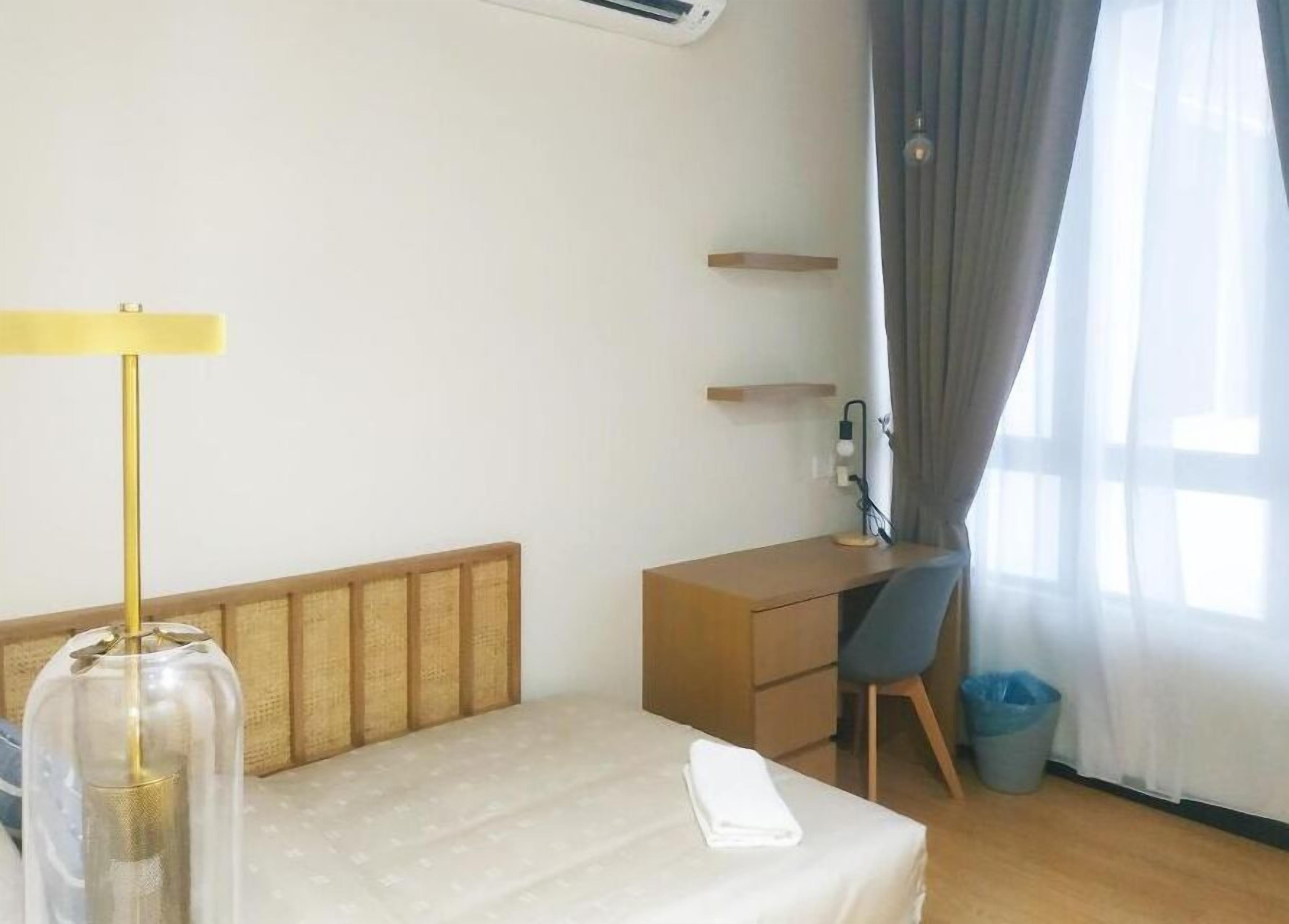 A hotel room with a bed, a desk, a chair, a lamp, a trash bin, and a window with curtains.