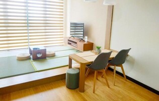 Una habitación con esteras de tatami, una mesa de madera, dos sillas, un televisor sobre un mueble bajo y una pequeña planta.