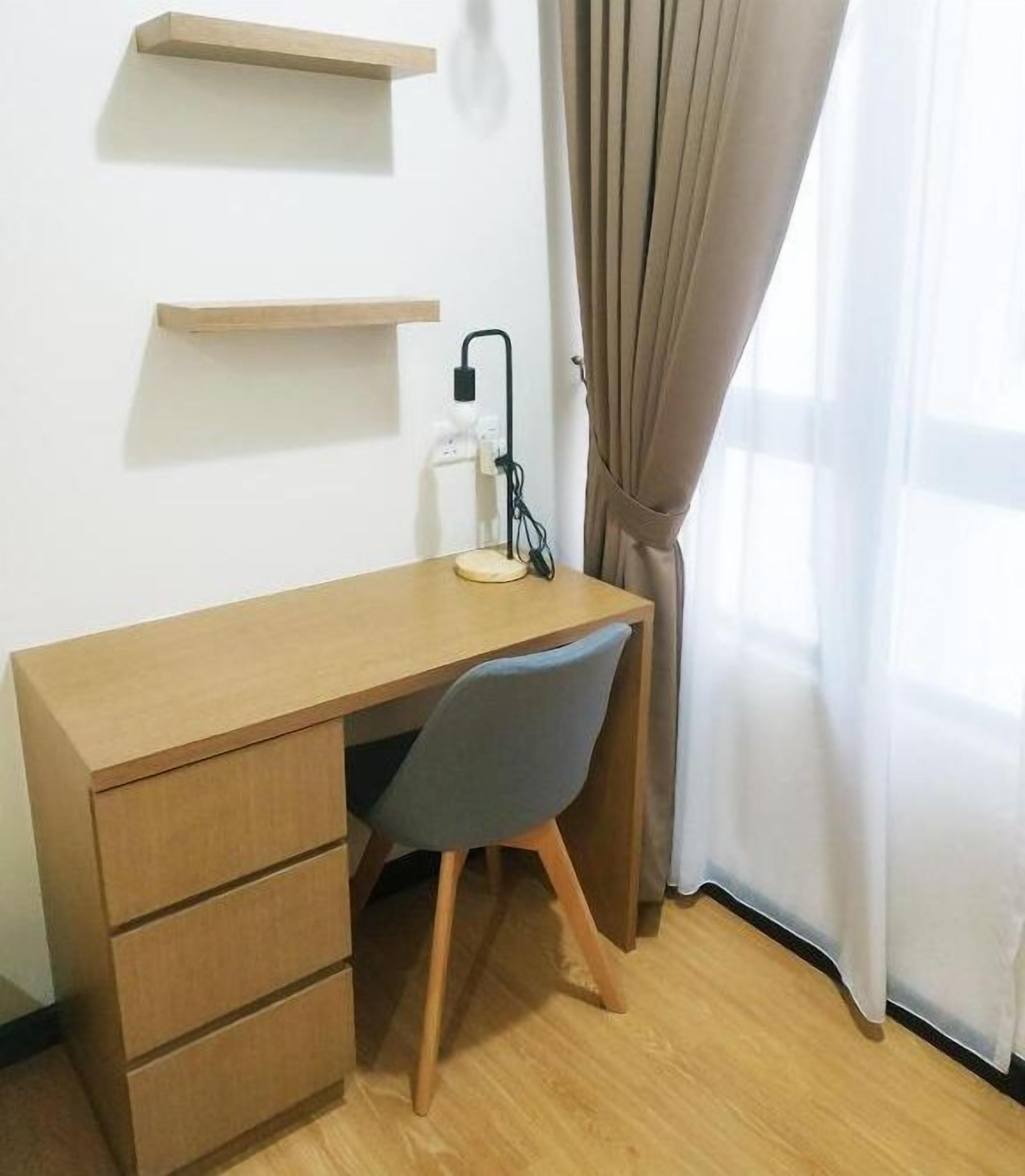 A wooden desk with a chair, a lamp, and two shelves against a white wall.