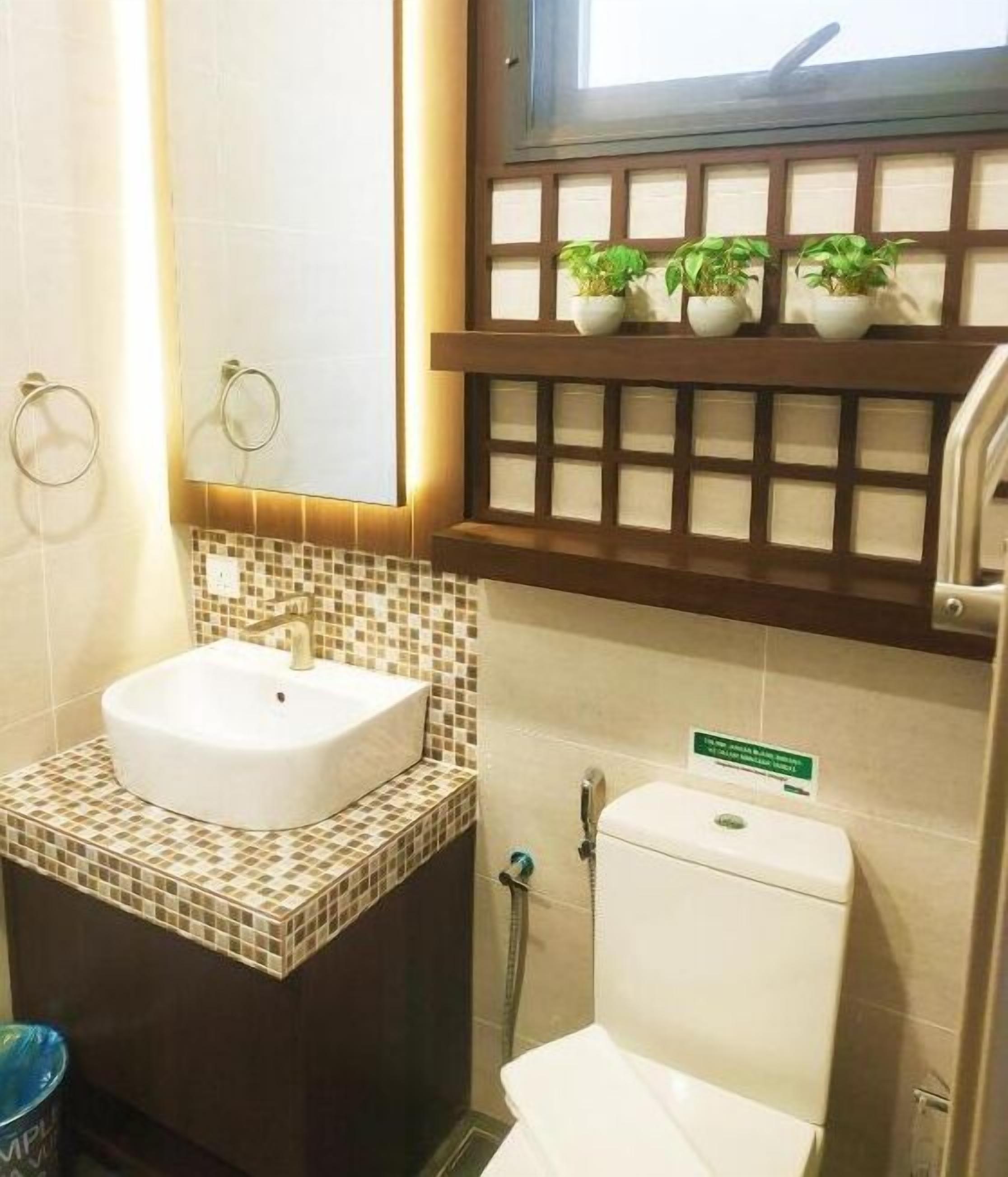 A bathroom with a white sink, a toilet, and a window above the toilet. There are three potted plants on a shelf above the window.
