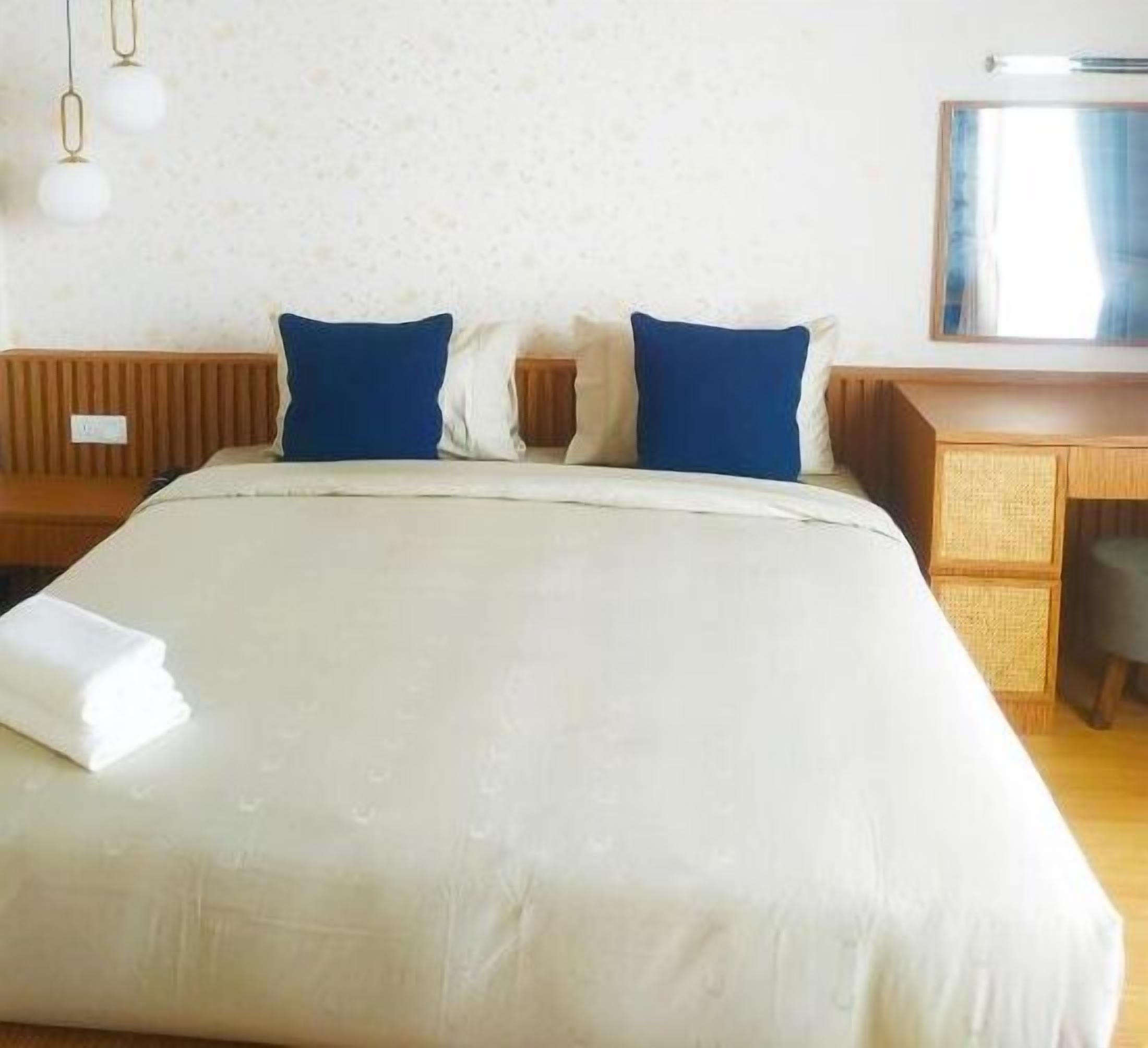 Une chambre d’hôtel avec un grand lit, deux oreillers bleus, une tête de lit en bois, un bureau avec une chaise et un miroir.