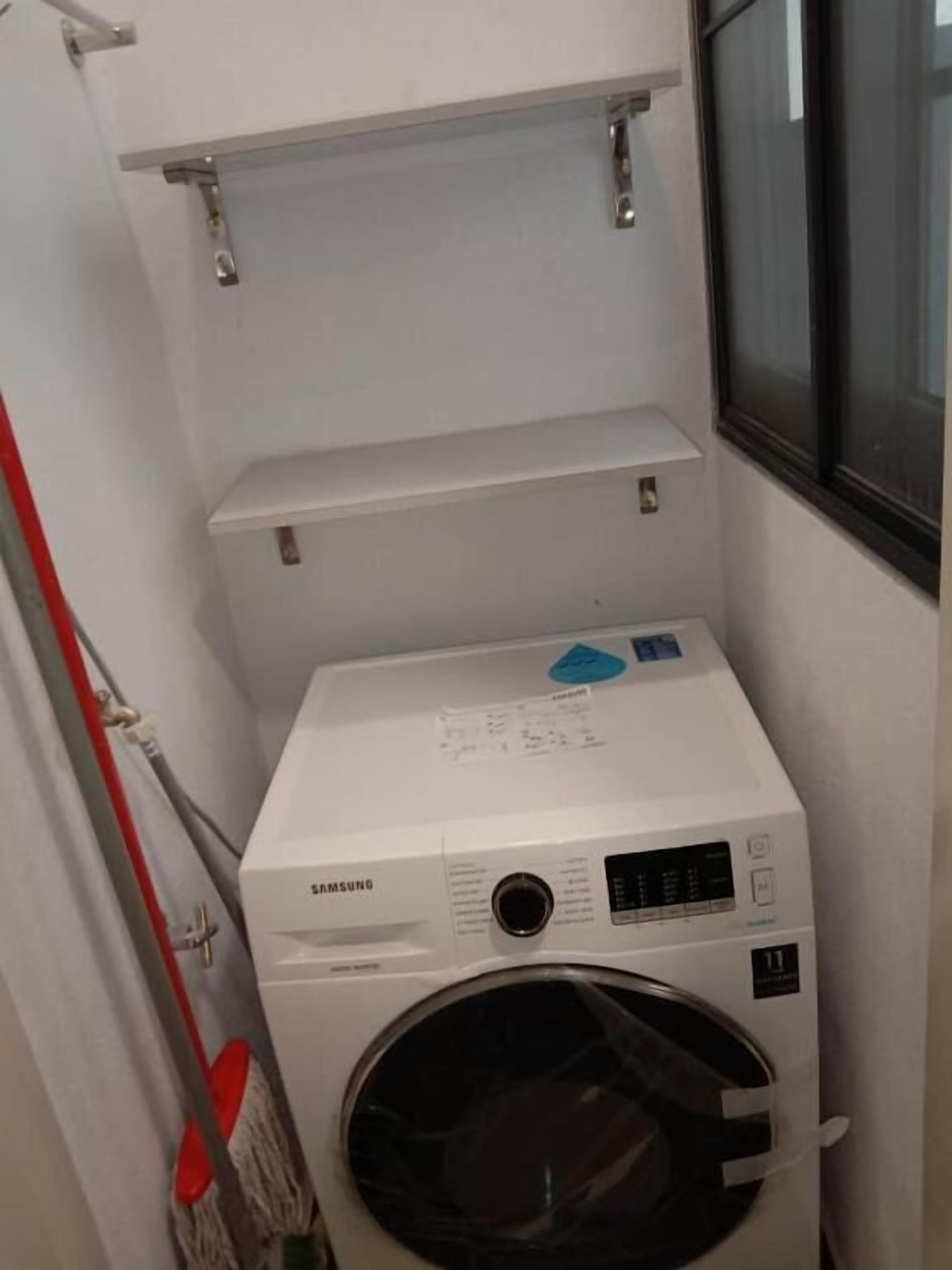 A compact Samsung washing machine in a small laundry room with a window and metal shelves.