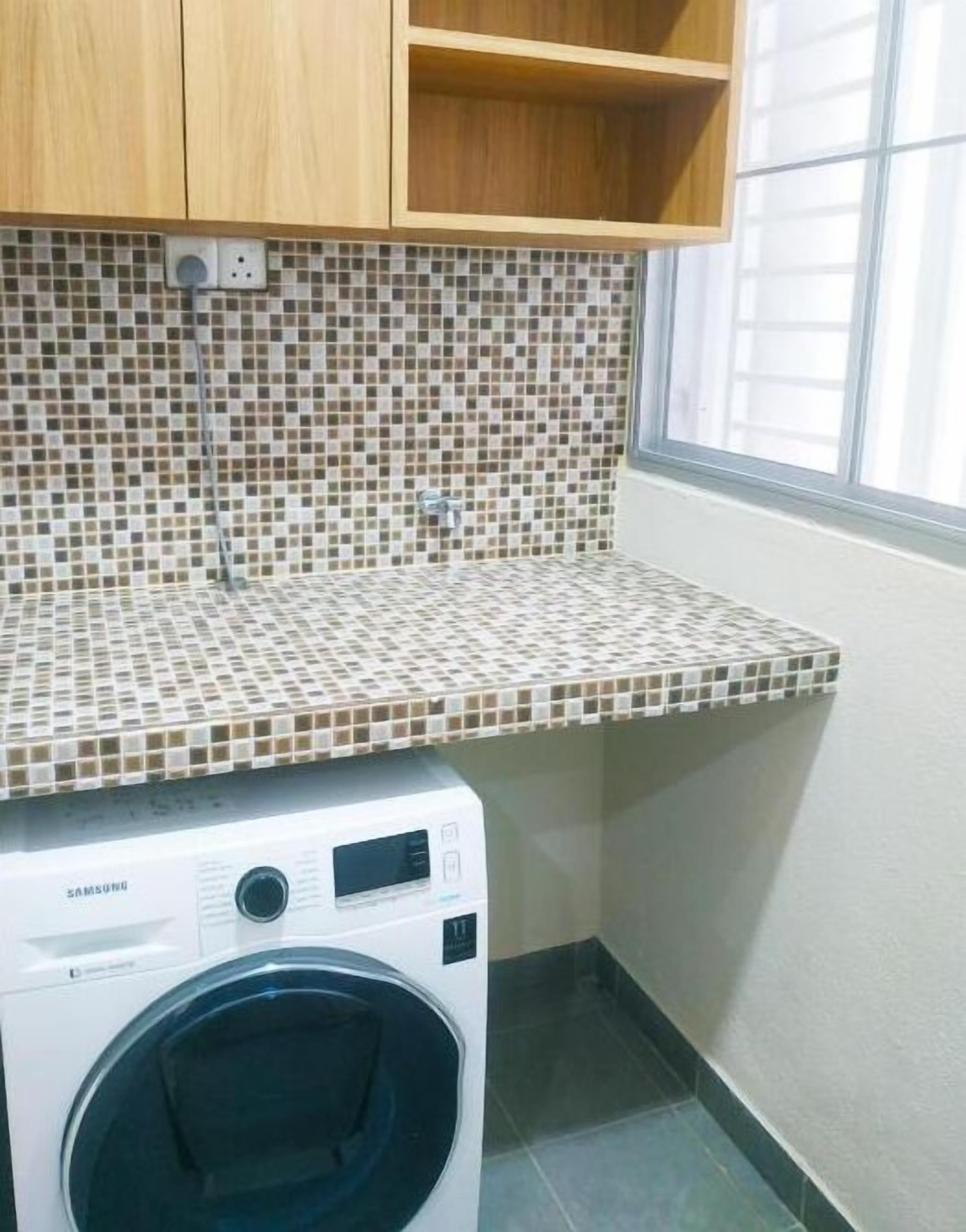 A laundry area with a washing machine, tiled backsplash, wooden cabinets, and a window.