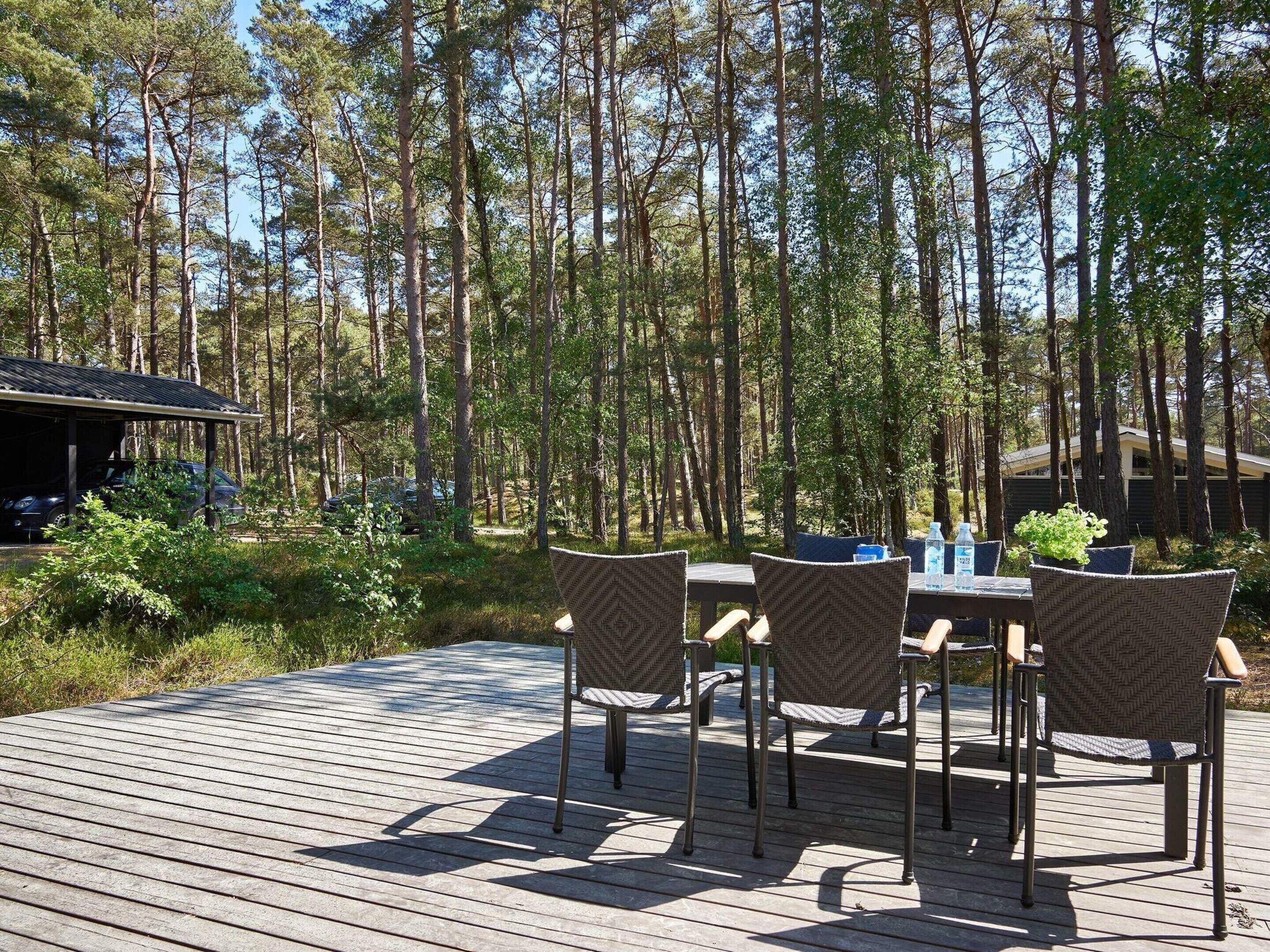 Una terraza de madera con cuatro sillas de mimbre y una mesa, rodeada de un bosque.
