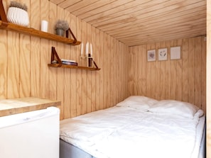 A bedroom with a wooden ceiling, a bed with white bedding, a small table with a white appliance, and wooden shelves with decorative items. - 4 Person Holiday Home in Bogense (Bogense)
