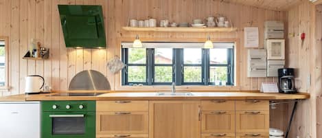 Une cuisine avec des meubles de rangement en bois, un poêle vert, une fenêtre avec des stores et une machine à café.