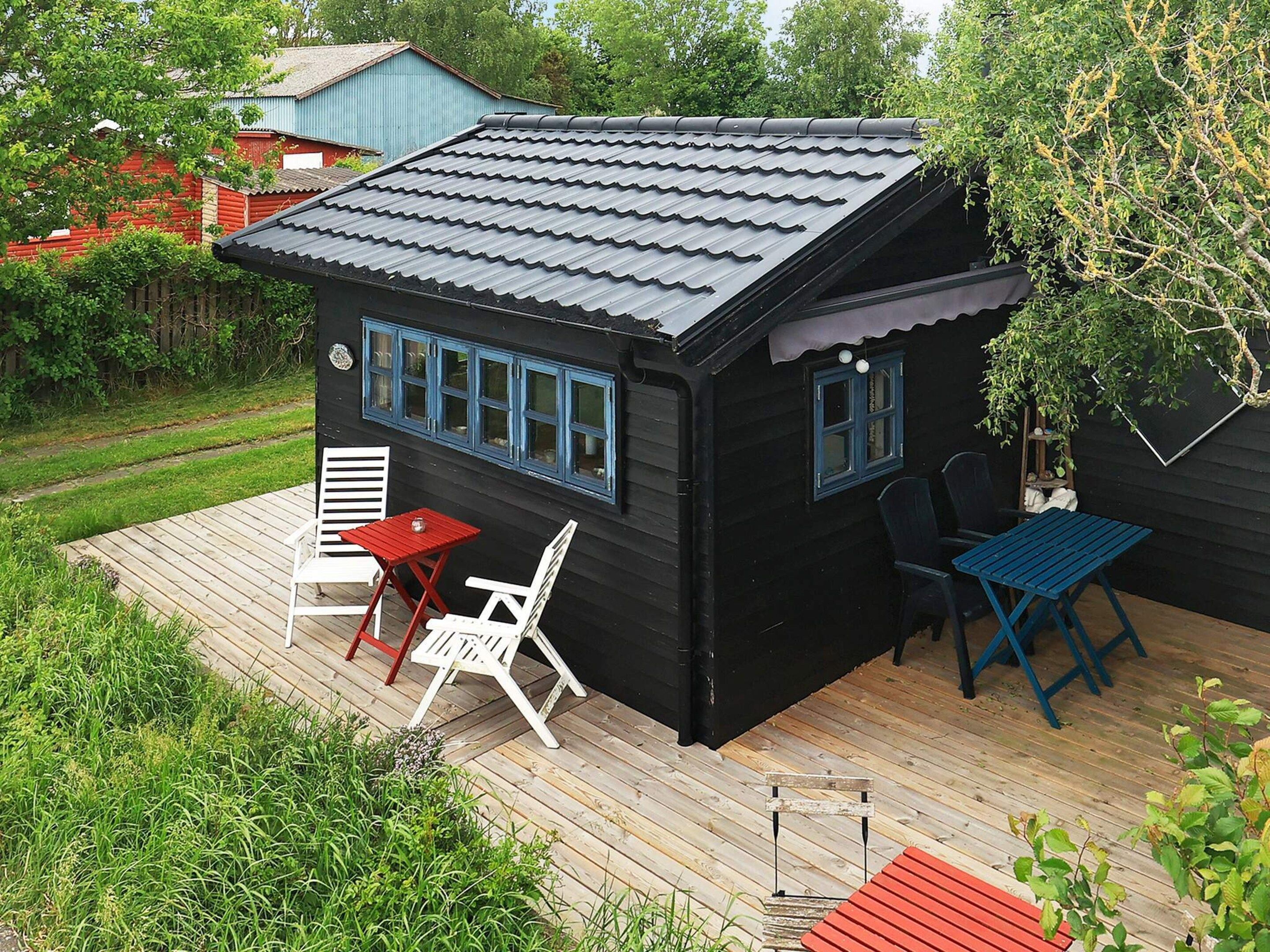 A small black cabin with a sloped roof, a deck with outdoor seating, and a view of the surrounding greenery.