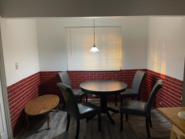 Habitación de esquina con una mesa redonda, cuatro sillas, una pequeña mesa de madera y una ventana con persianas.