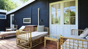 A wooden deck with wicker furniture, including a bed and chairs, and a large window with a view of greenery. - 4 Star Holiday Home in Vejby (Vejby)