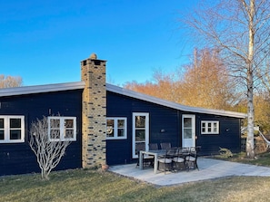 A single-story house with a chimney, a patio with a table and chairs, and a small tree in the front yard. - 4 Star Holiday Home in Vejby (Vejby)