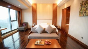 Une chambre d’hôtel moderne avec un grand lit, un canapé, une table basse en bois sur laquelle sont disposés des fruits et un plateau, et une vue sur la ville grâce à de grandes fenêtres.