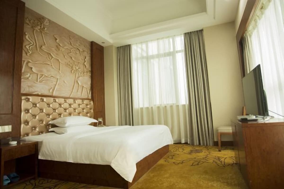 A hotel room with a large bed, a wooden headboard with a carved design, a television on a wooden desk, and a window with sheer curtains.