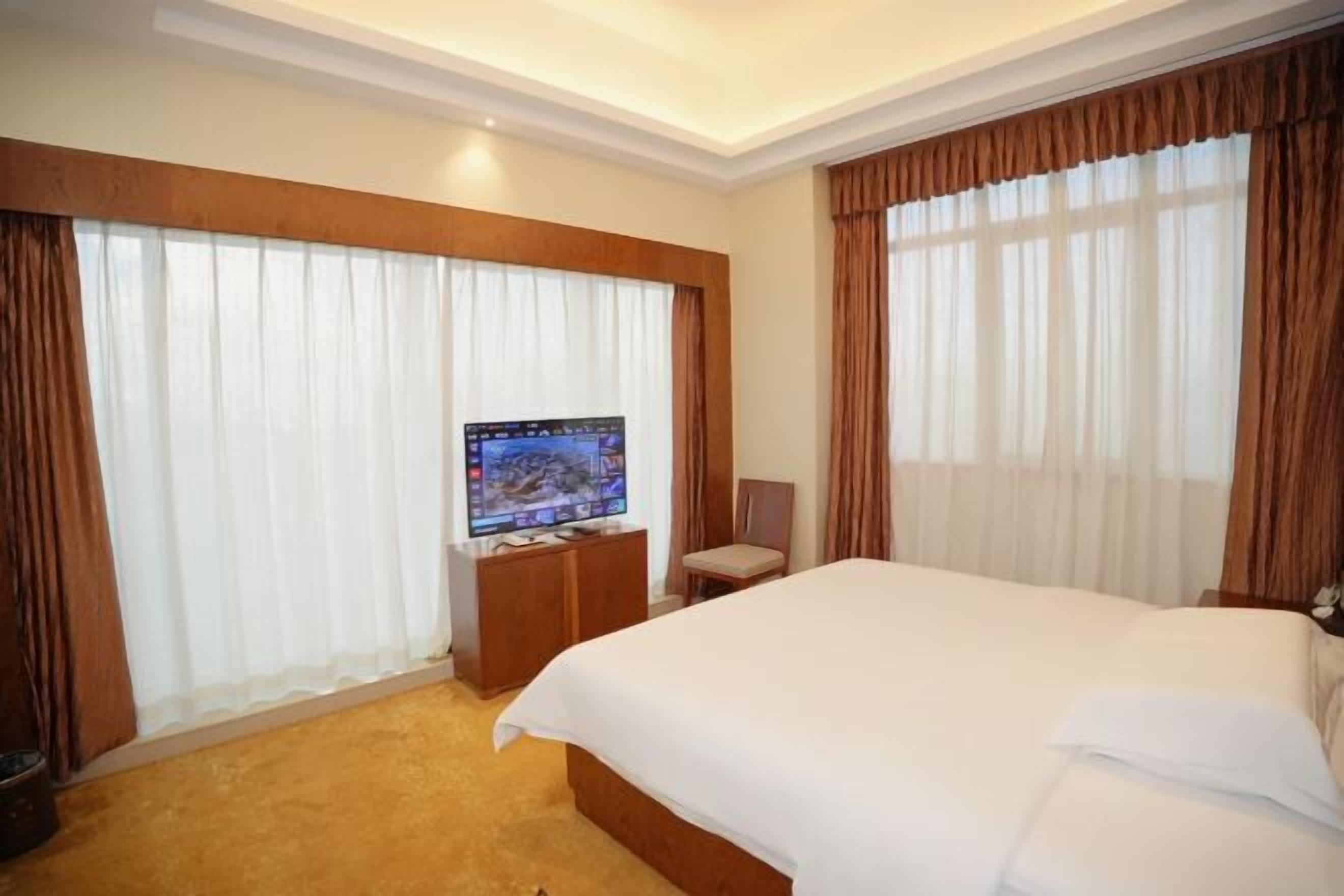 A hotel room with a large bed, a television on a wooden stand, and curtains on the windows.