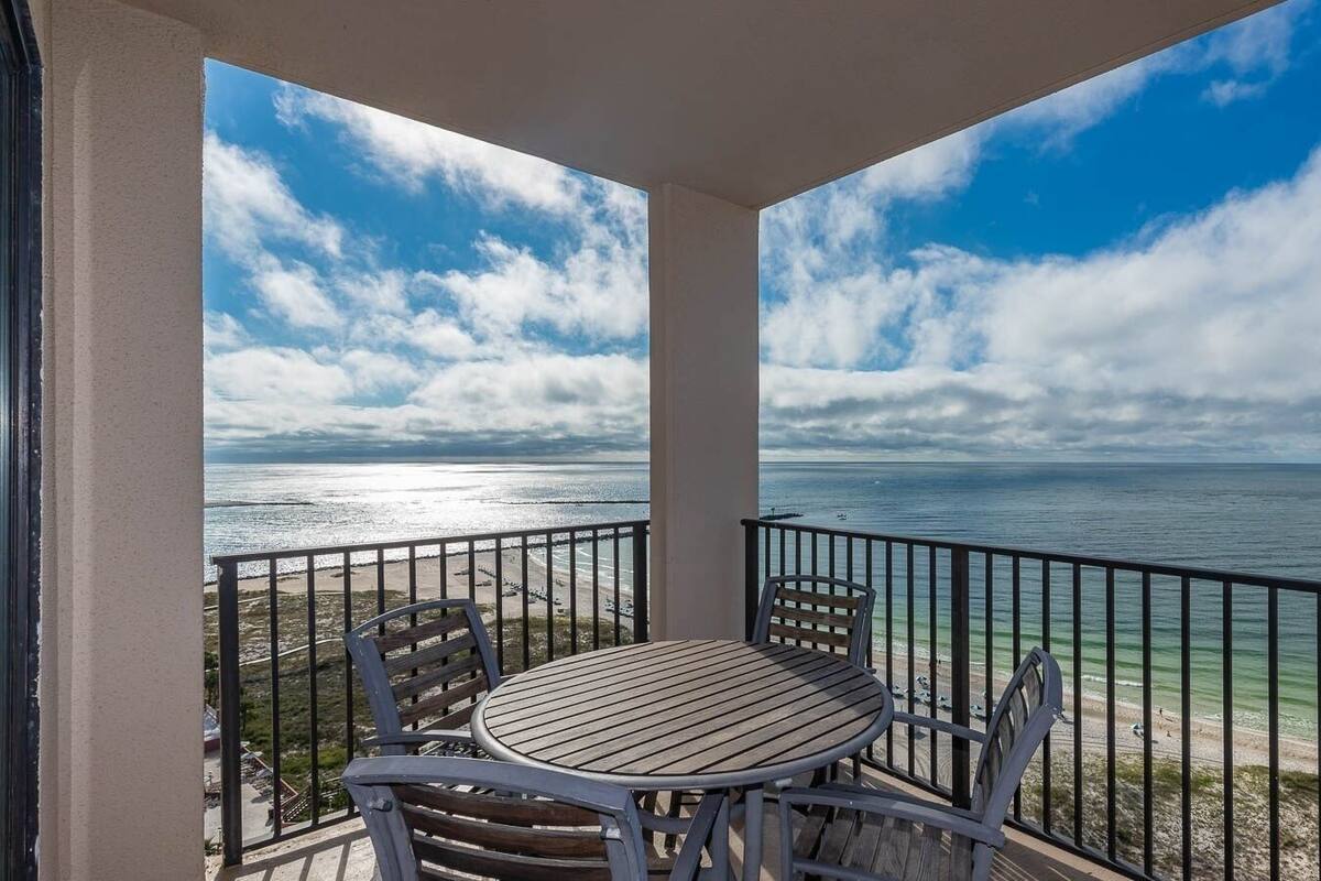 Een balkon met een ronde houten tafel en stoelen, met uitzicht op een strand en heldere lucht boven een zee.