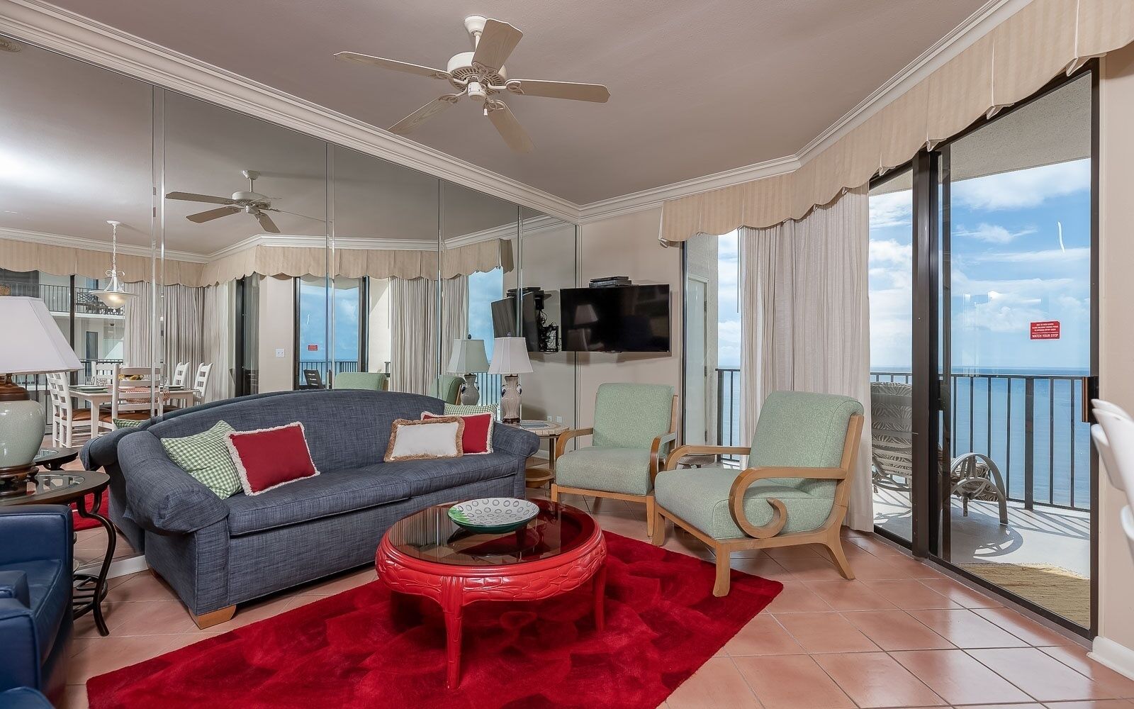 Un salon spacieux avec un canapé gris, des coussins rouges et verts, une table basse rouge et un balcon offrant une vue sur la mer.