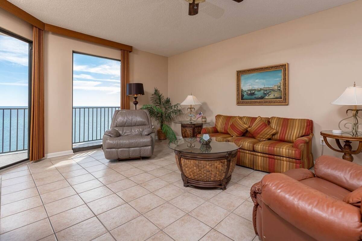 Een ruime woonkamer met een leren fauteuil, een gestreepte bank en een glazen salontafel. Er is een balkon met uitzicht op zee.