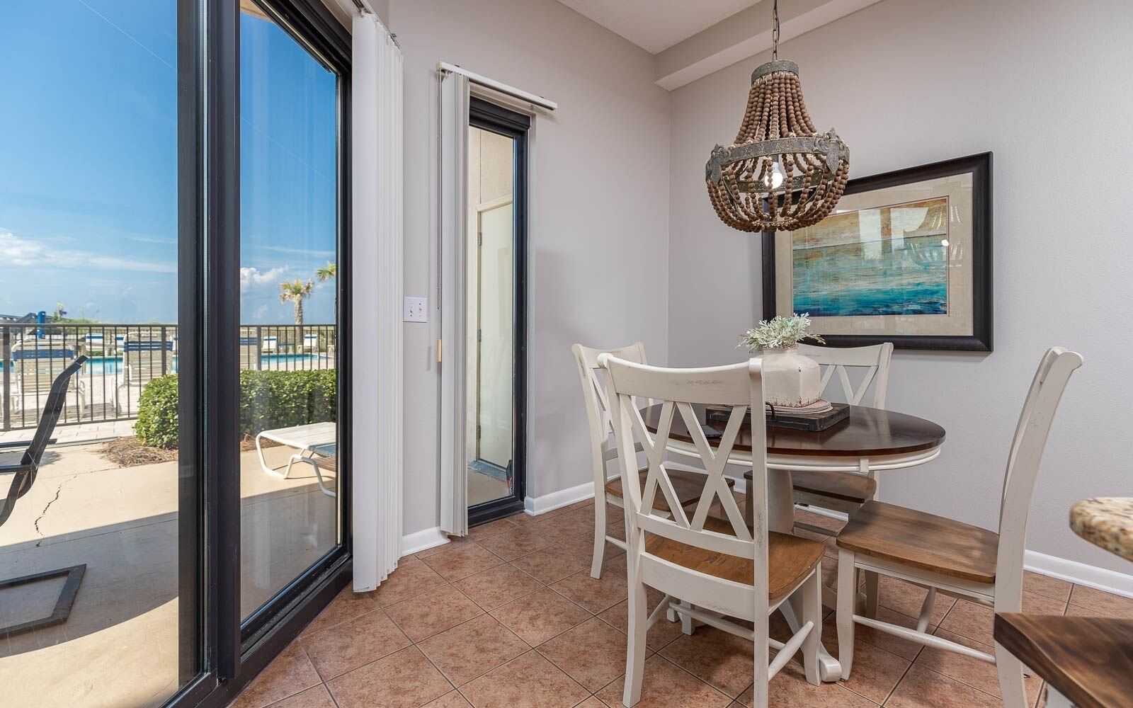 A dining area with a table and chairs, a chandelier, a painting on the wall, and a view of a pool and palm trees through a glass door.