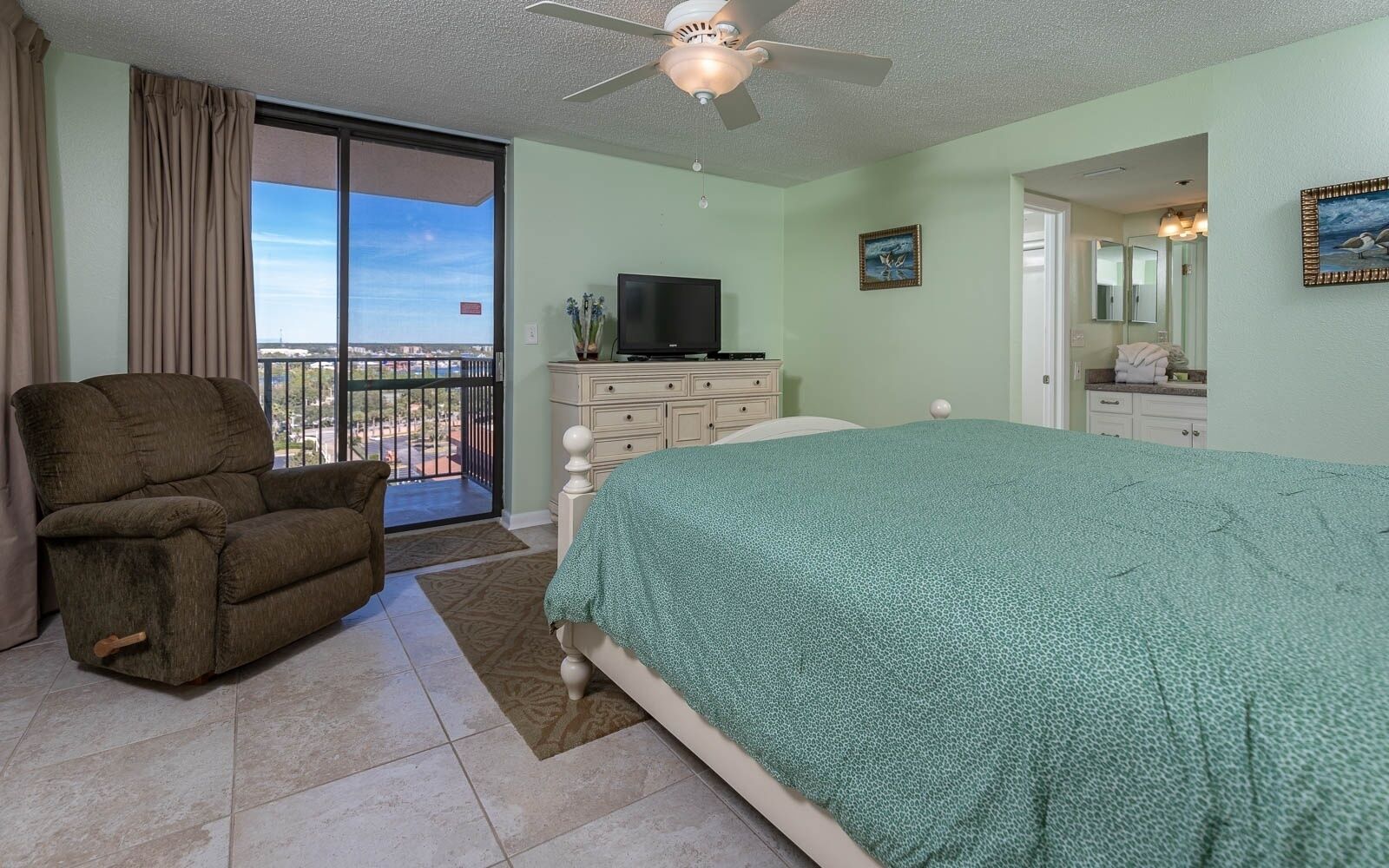 Un dormitorio con cama, una silla, tocador, televisor y balcón con vistas.