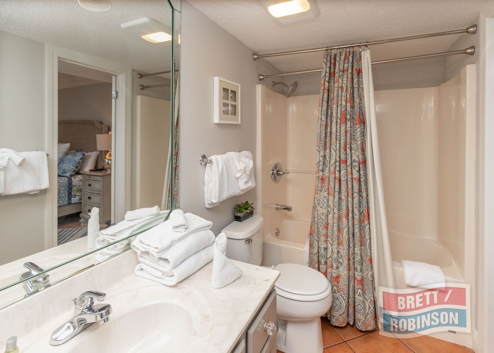 Une salle de bain avec un lavabo blanc, des toilettes, une douche avec un rideau à motifs et un miroir. Des serviettes sont pliées sur le lavabo et une petite plante est posée sur les toilettes.