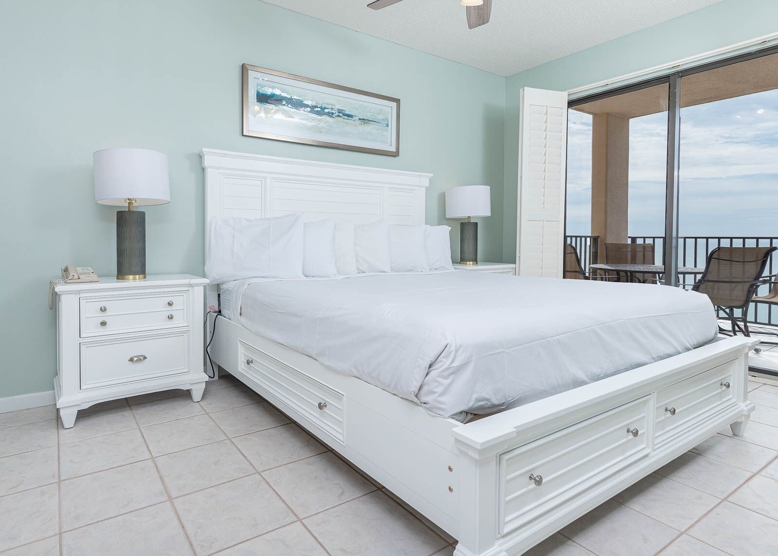 A bedroom with a large bed, two bedside tables with lamps, a framed picture on the wall, and a balcony with chairs.