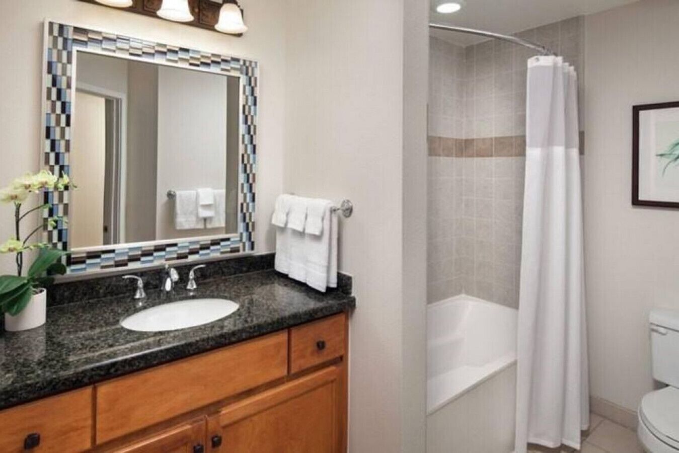 Une salle de bain avec un lavabo sur un plan de travail en granit, des meubles en bois, un grand miroir orné d’une bordure en mosaïque, un rideau de douche blanc et un tableau encadré accroché au mur.