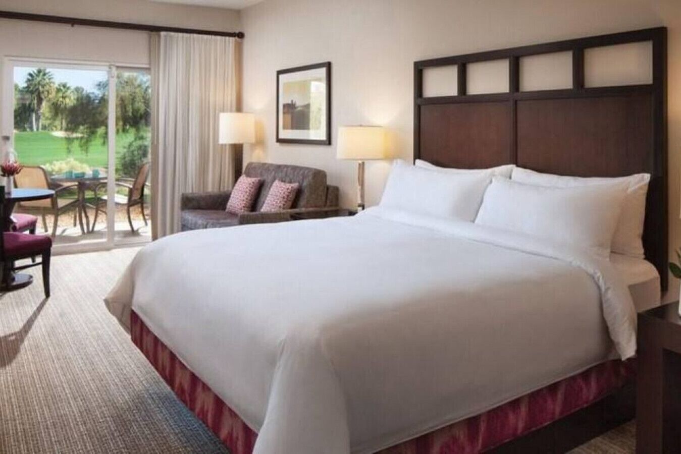 Ein Hotelzimmer mit einem großen Bett, einem Schreibtisch mit Stuhl, einer Lampe und Blick auf einen Golfplatz durch das Fenster.