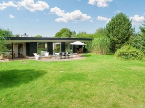 Et moderne hus med en terrasse, udendørs møbler og et stort træ i baggrunden.