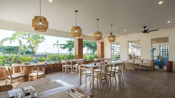 A spacious dining area with wooden tables and chairs, hanging rattan chandeliers, and large windows offering a view of the outdoors.