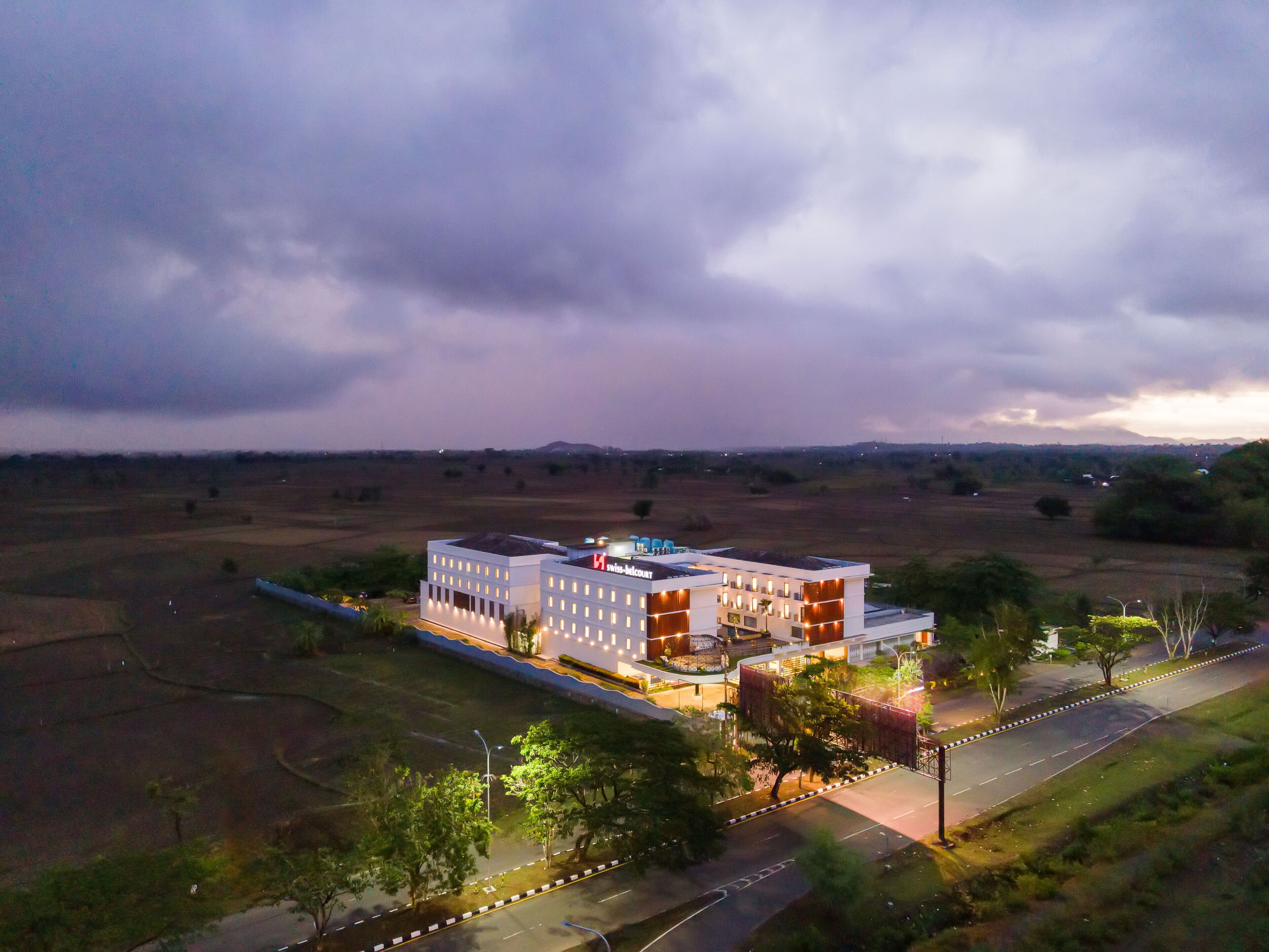 Un hotel moderno con techo plano y varios pisos, rodeado de campos abiertos y algunos árboles.