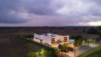 A modern hotel with a flat roof and multiple floors, surrounded by open fields and a few trees.
