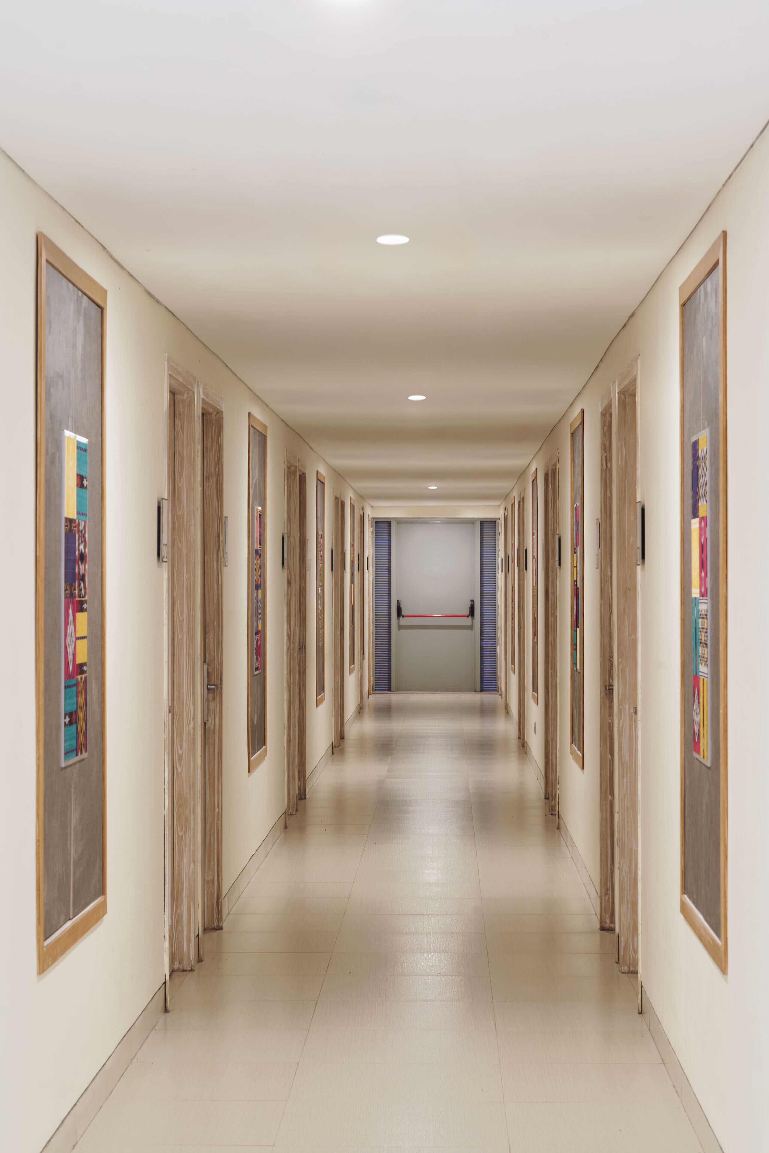 A long, narrow hallway with doors on both sides, leading to a room with a large window and a red barrier.