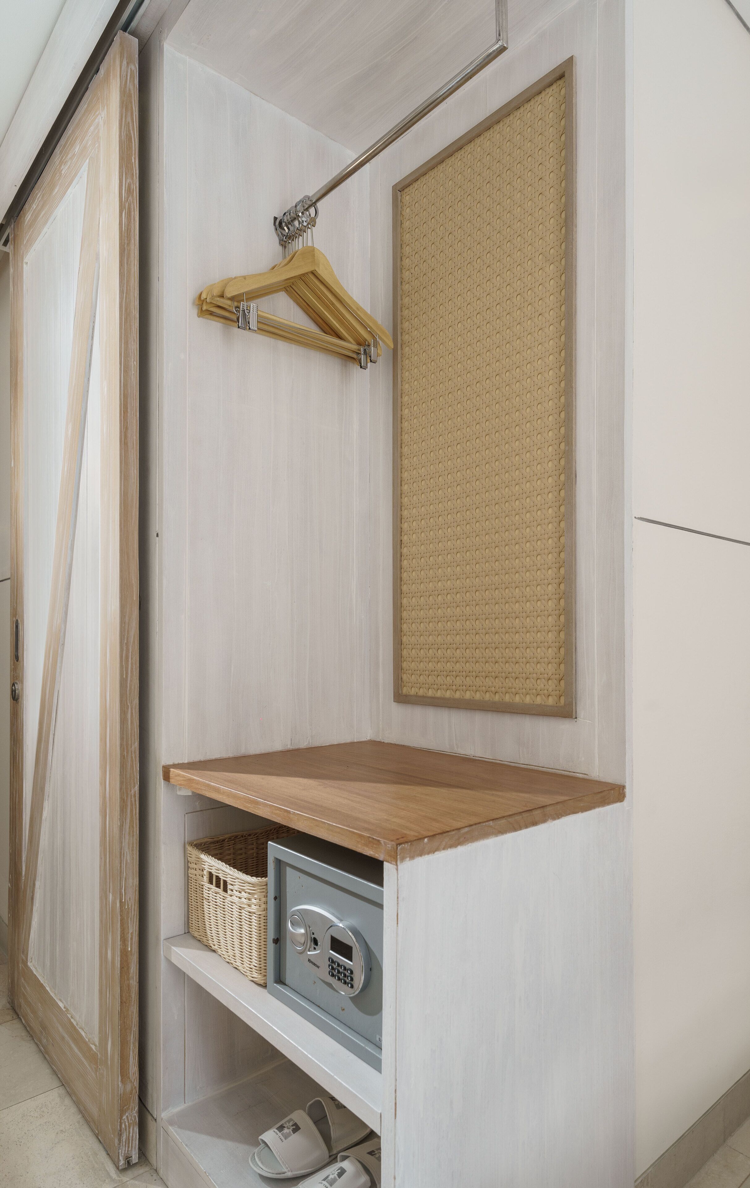 A room with a wooden wardrobe, a hanging rack, a woven bulletin board, a small wooden shelf with a basket, and a safe.