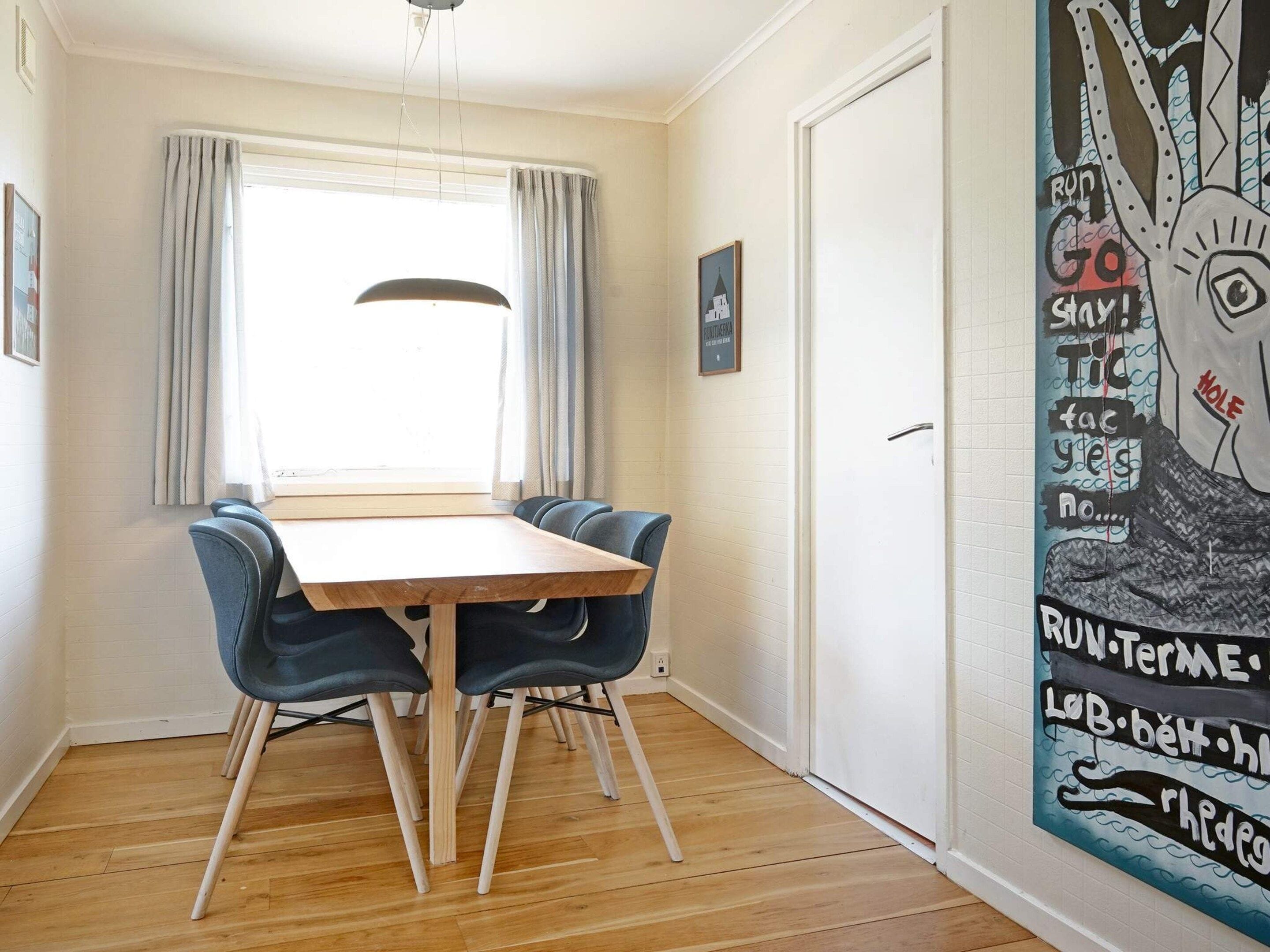A dining area with a wooden table and blue chairs, a window with curtains, and a wall with a large abstract painting.