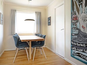 Een eetgedeelte met een houten tafel en blauwe stoelen, een raam met gordijnen, en een muur met een groot abstract schilderij.