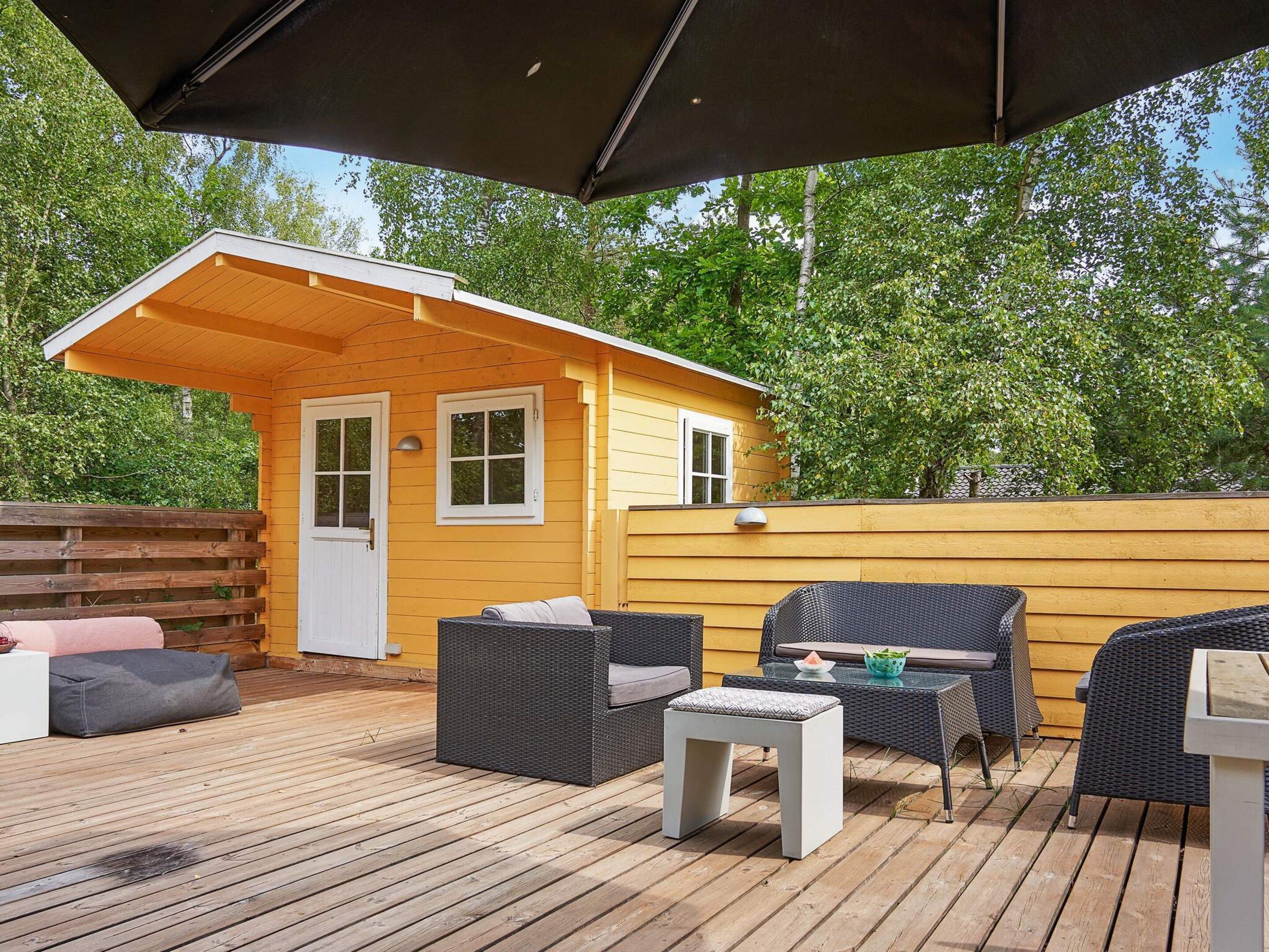 Une terrasse en bois avec des meubles d’extérieur, un petit bâtiment jaune avec une porte et un grand parasol.