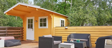 Una terraza de madera con mobiliario exterior, un pequeño edificio amarillo con una puerta y un paraguas grande.