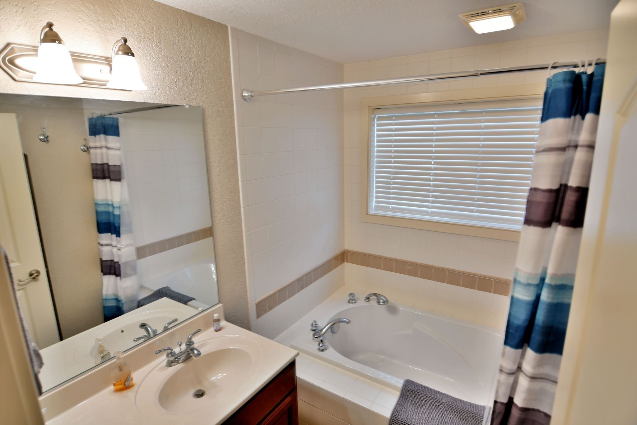 Une salle de bain avec un double lavabo, une baignoire, une fenêtre avec des stores et un rideau de douche.