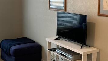 Una habitación con televisión de pantalla plana sobre un soporte blanco, una ventana con persianas, un otomano de color oscuro y piso alfombrado.