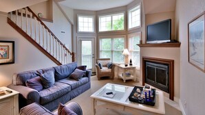 Una sala de estar con escalera, chimenea, televisor y un rincón con ventanas.
