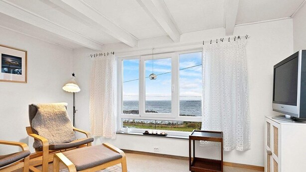 A room with a wooden chair, a small table, a TV on a white cabinet, and a window with a view of the sea.