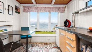 A compact kitchen with a view of the sea, a dining area with a blue chair, and patterned floor tiles.