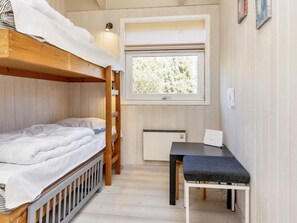 A compact room with bunk beds, a small table with a chair, a window with blinds, and a wall-mounted heater.