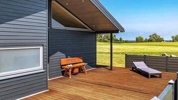 A modern outdoor deck with a wooden bench and a lounge chair, a covered area, and a view of a grassy field.