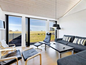 A modern living room with a sofa, a coffee table, a chair, and a TV. There are large windows offering a view of the outdoors. - 5 Star Holiday Home in Rudkobing (Rudkobing)