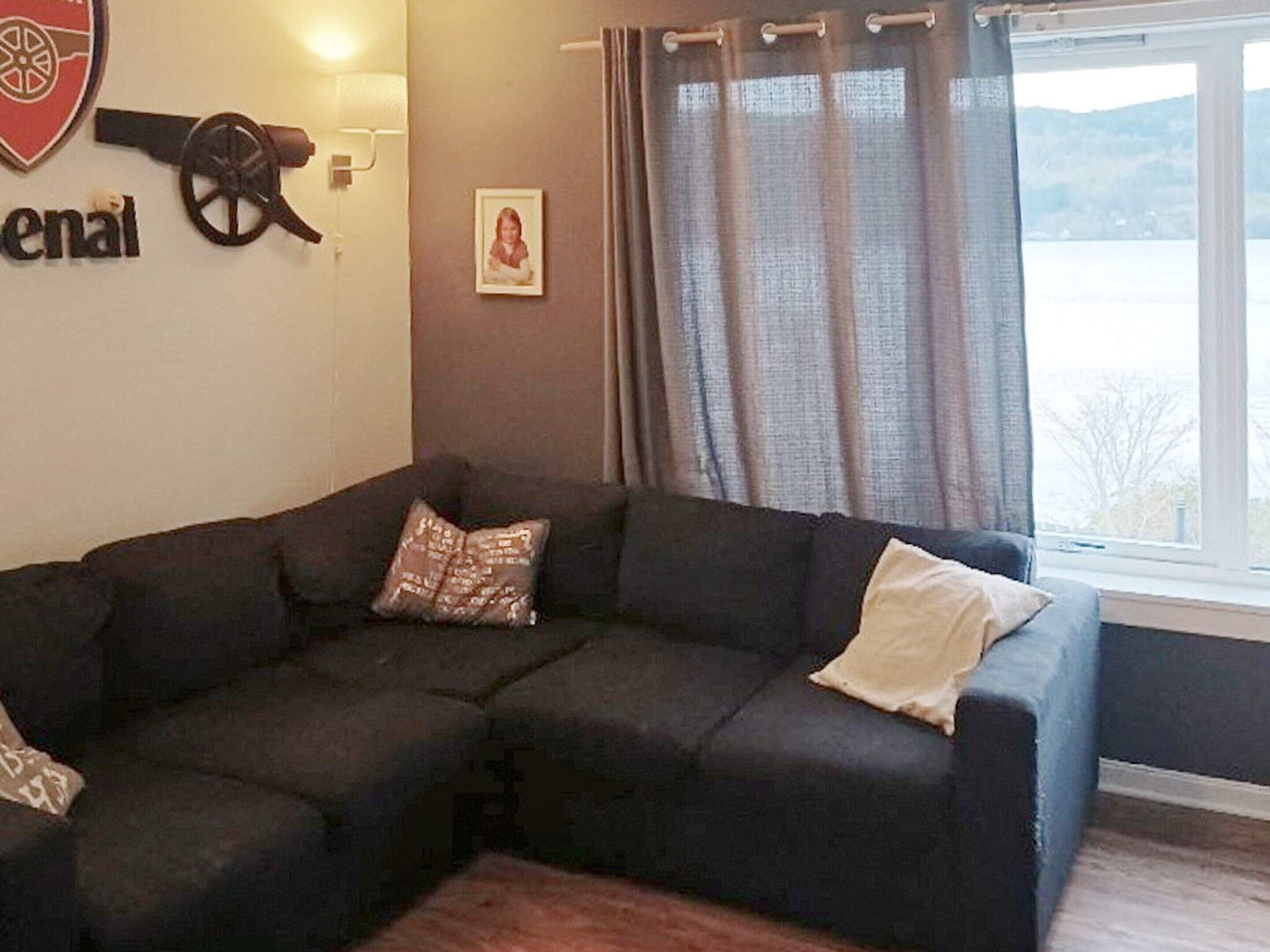 A living room with a sectional sofa, a wall-mounted cannon, a framed picture, and a view of a body of water through a window with curtains.