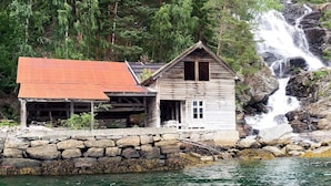 Une cabane rustique à toit rouge, située au bord d’une côte rocheuse, avec une cascade en arrière-plan.