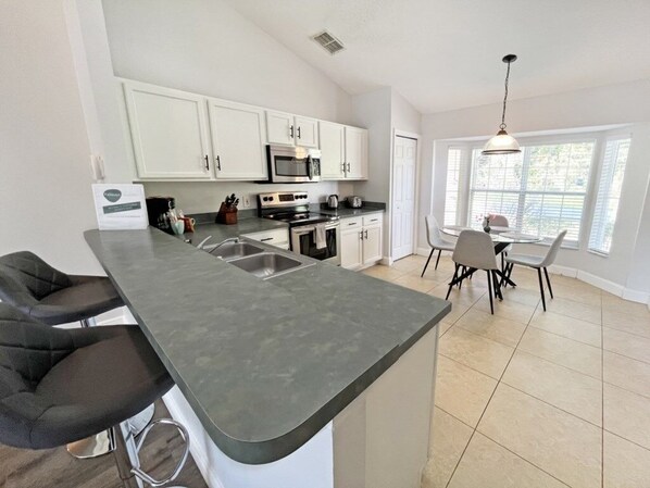 Una cocina moderna con armarios blancos, una isla grande con taburetes de bar, un comedor con mesa y sillas, y un ventanal que proporciona luz natural.