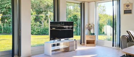A modern living room with a flat-screen TV, wooden flooring, and large windows offering a view of a garden.