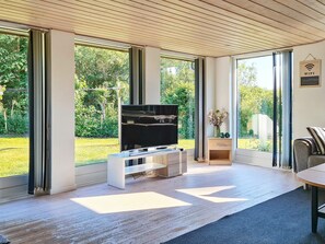 A modern living room with a flat-screen TV, wooden flooring, and large windows offering a view of a garden. - 6 Person Holiday Home in Hals (Hals)
