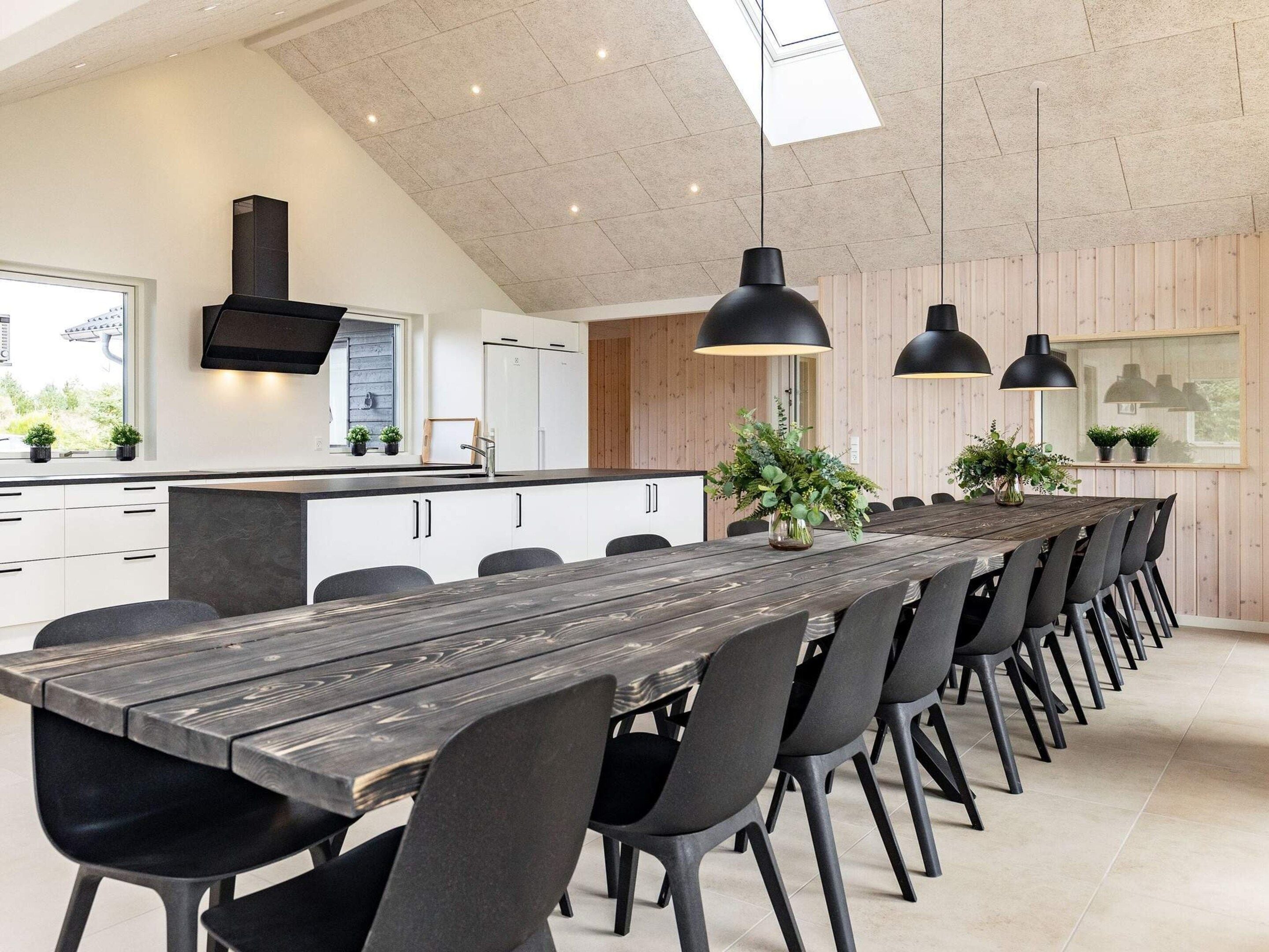 Een moderne keuken met een lange eettafel, zwarte stoelen, hanglampen en houten keukenkasten.